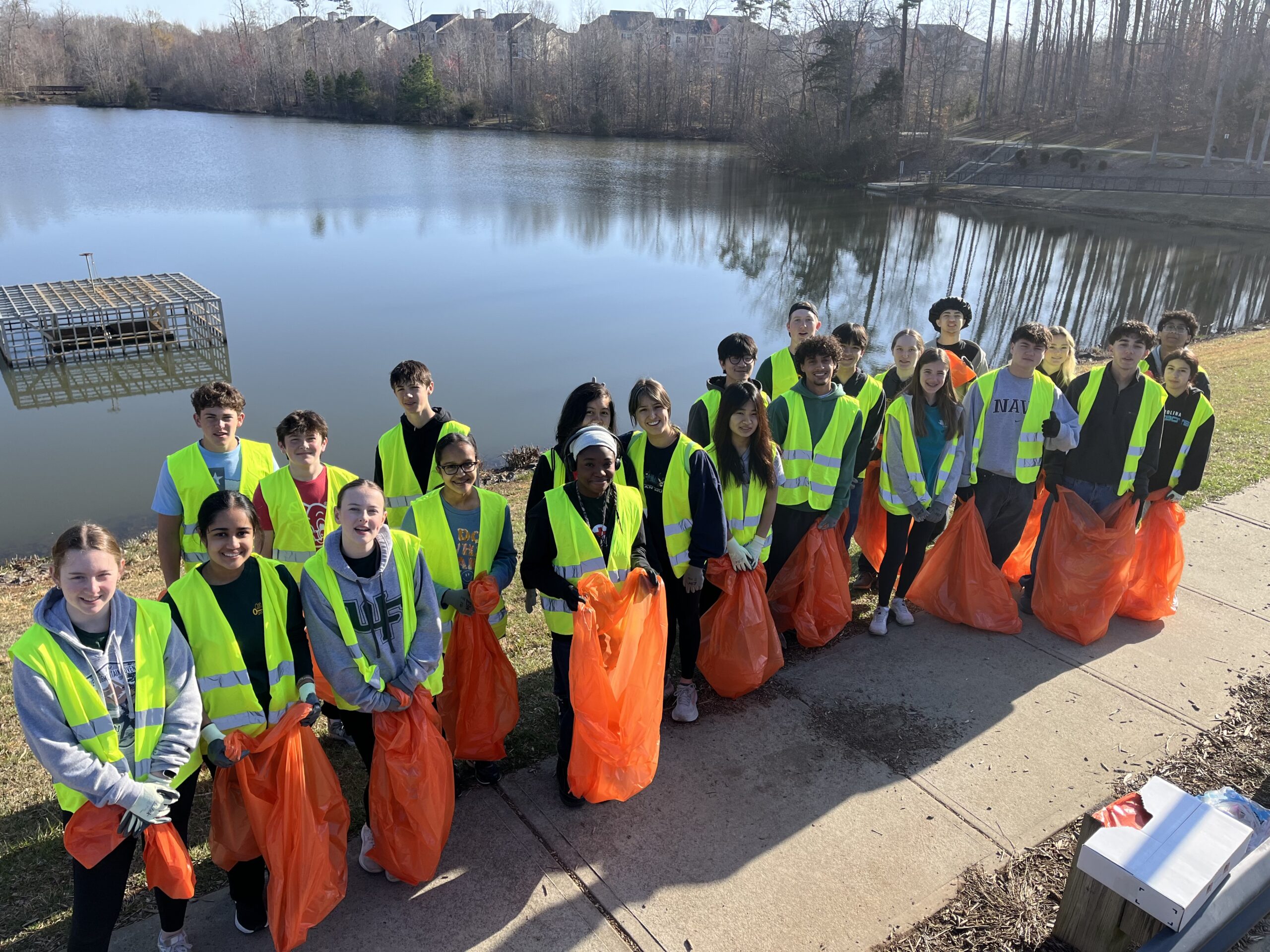 Clemmons_Cleanup2 (1) A group of students in yellow safety vests pose in front of a lake with orange trash bags
