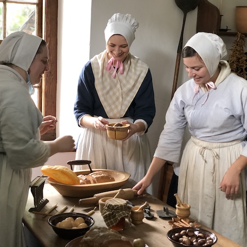 Three women around a table of bread in historically accurate Moravian clothing